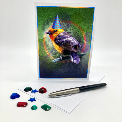 Photo of a greeting card posed with plastic rhinestones and an ink pen. The card features a flame-colored tanager in a blue conical party hat against a green and purple background.