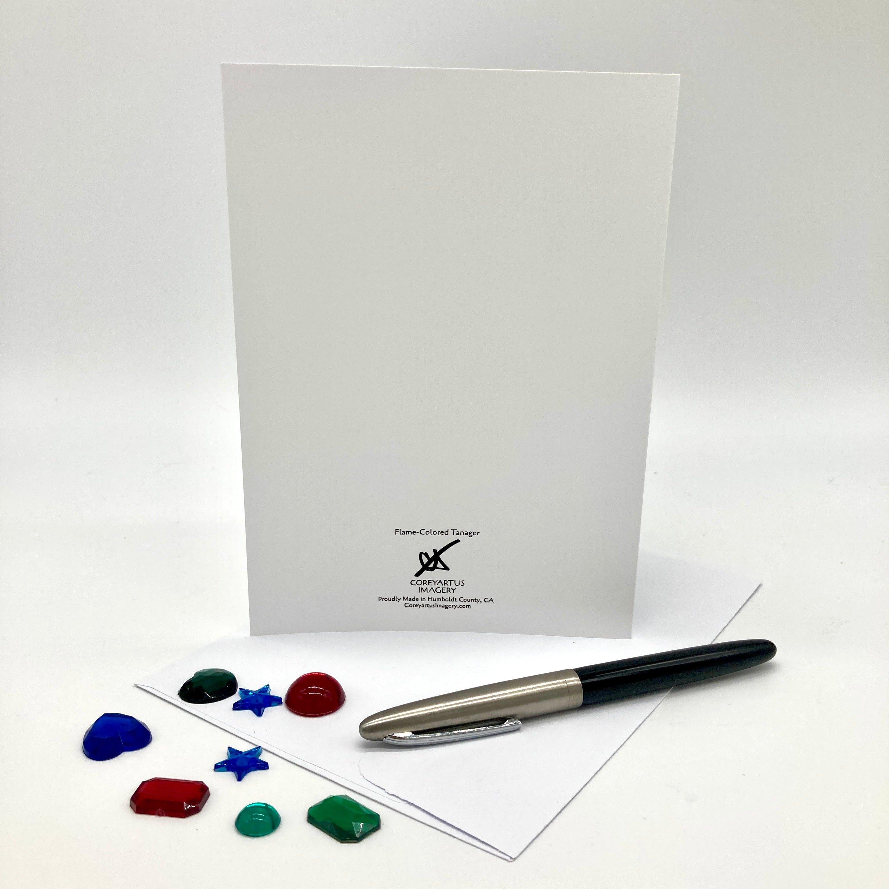 A white stationery set featuring a folded white card with black printed branding at the bottom, a black and silver pen resting on a flat white sheet, and several small colorful decorative stones in blue, green, red, and black arranged in front, all photographed on a clean white studio background.