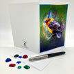 Photo of a splayed open greeting card posed with plastic rhinestones and an ink pen. The card features a flame-colored tanager in a blue conical party hat against a green and purple background.