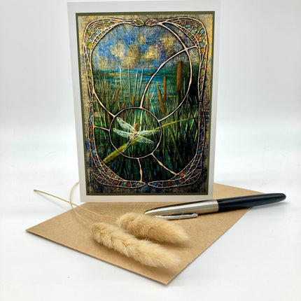A vertical art print or card featuring an abstract stained-glass-style landscape with a dragonfly, displayed upright on a kraft envelope with a pen and dried grass stems on a white background.