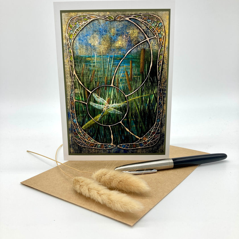 A vertical art print or card featuring an abstract stained-glass-style landscape with a dragonfly, displayed upright on a kraft envelope with a pen and dried grass stems on a white background.