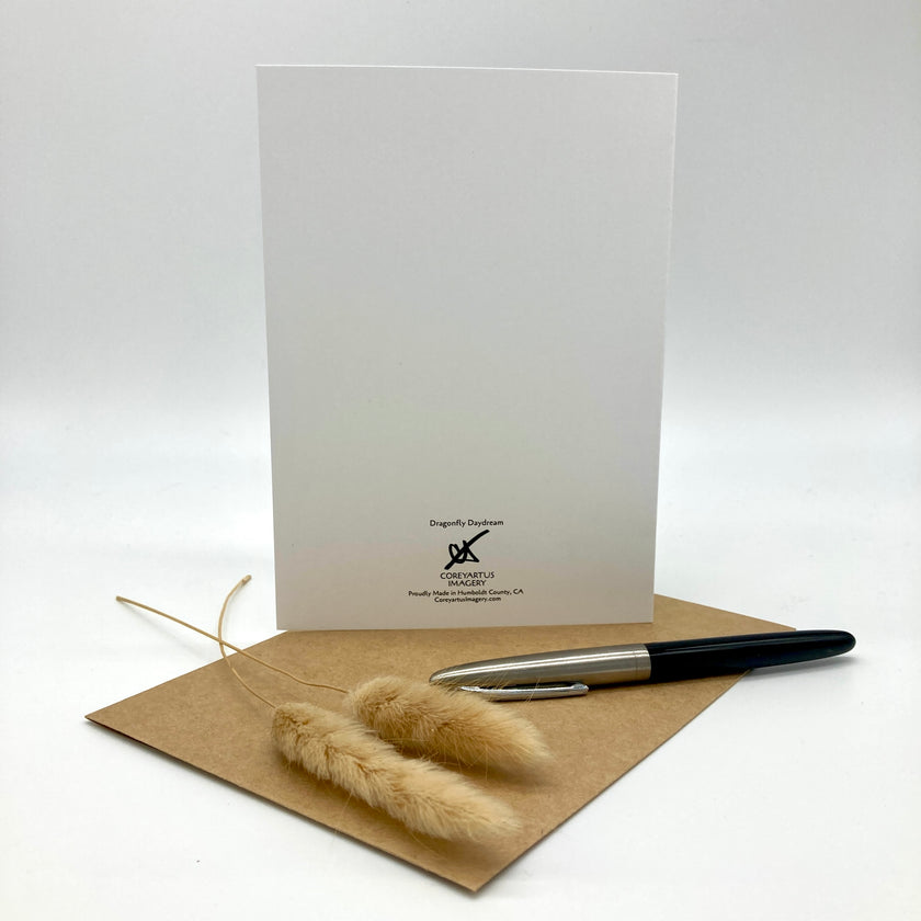 A white greeting card with black printed branding stands upright on a brown kraft envelope, with a black and silver pen and two dried pampas grass stems resting in front on a light gray studio background.