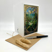 A white greeting card with a stained-glass style illustration of a lily of the valley, displayed on a white background with a brown kraft envelope, a black and silver pen, and dried bunny tail grass.