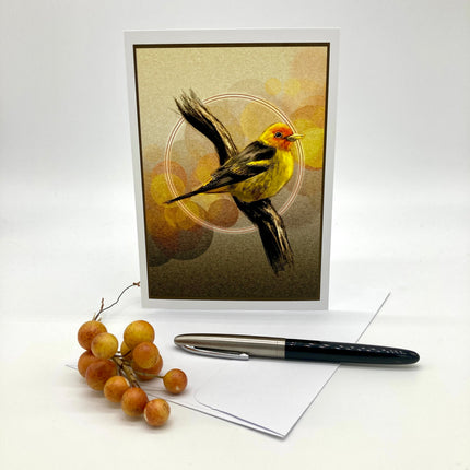 A vertical greeting card featuring a painted yellow bird with an orange head on a textured beige and brown background, displayed standing upright with a white envelope and a black pen on a white surface. A small cluster of brown grapes is placed beside the card.