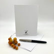 A white greeting card with a black printed logo and text at the bottom stands upright on a white surface, with a white envelope and a black-and-silver pen placed in front. A small decorative cluster of amber-orange berries rests on the left side.