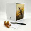A white greeting card with a printed illustration of a yellow bird on the front, displayed with an envelope and a black pen on a white background. The card shows the brand name 'The Art of X' printed near the bottom left.