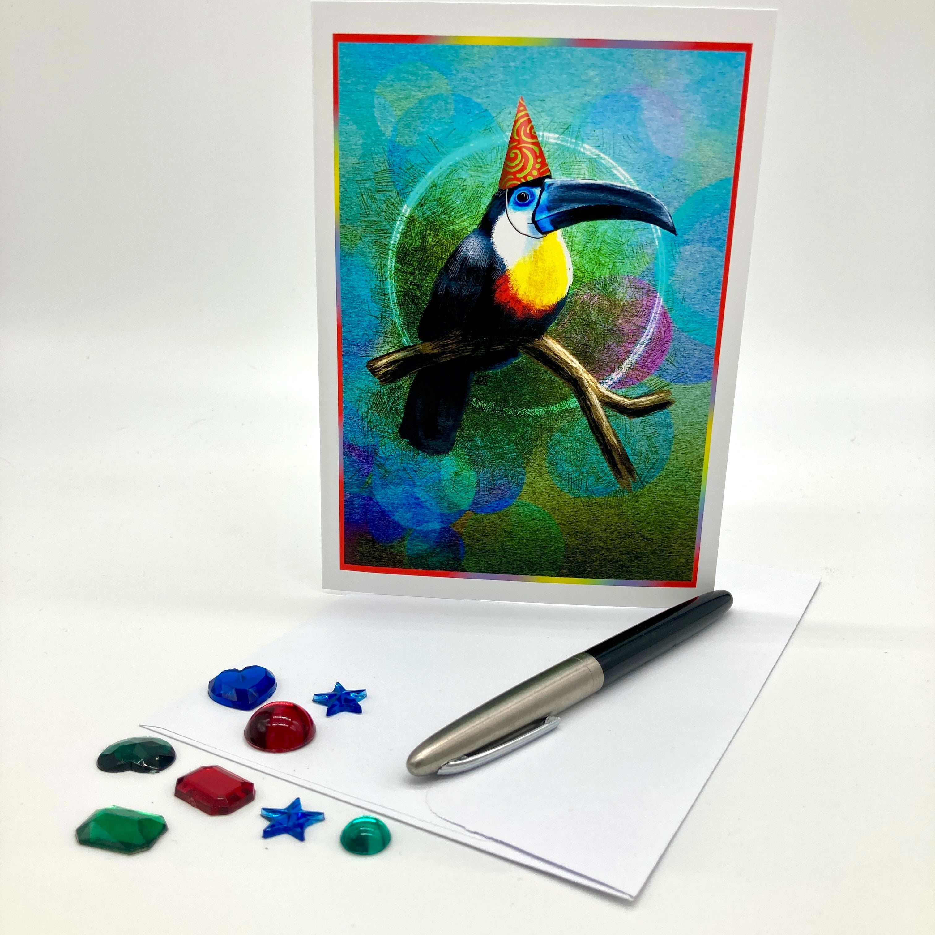 A greeting card featuring a colorful toucan perched on a branch against an abstract blue and green background, displayed upright with an envelope, a pen, and small decorative stones on a white studio surface.