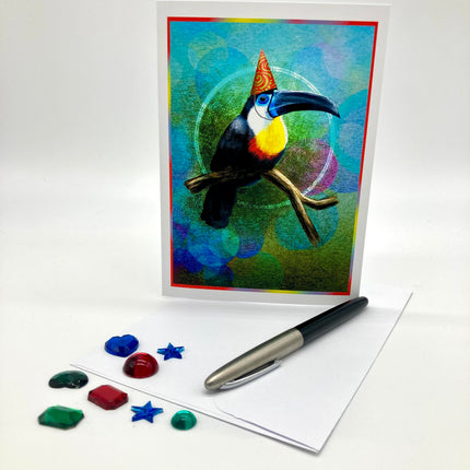 A greeting card featuring a colorful toucan perched on a branch against an abstract blue and green background, displayed upright with an envelope, a pen, and small decorative stones on a white studio surface.