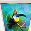 A colorful art print featuring a toucan perched on a branch, wearing a red and orange party hat. The background is a vibrant mix of blue, green, and purple textured circles, framed by a white border with red and blue lines.
