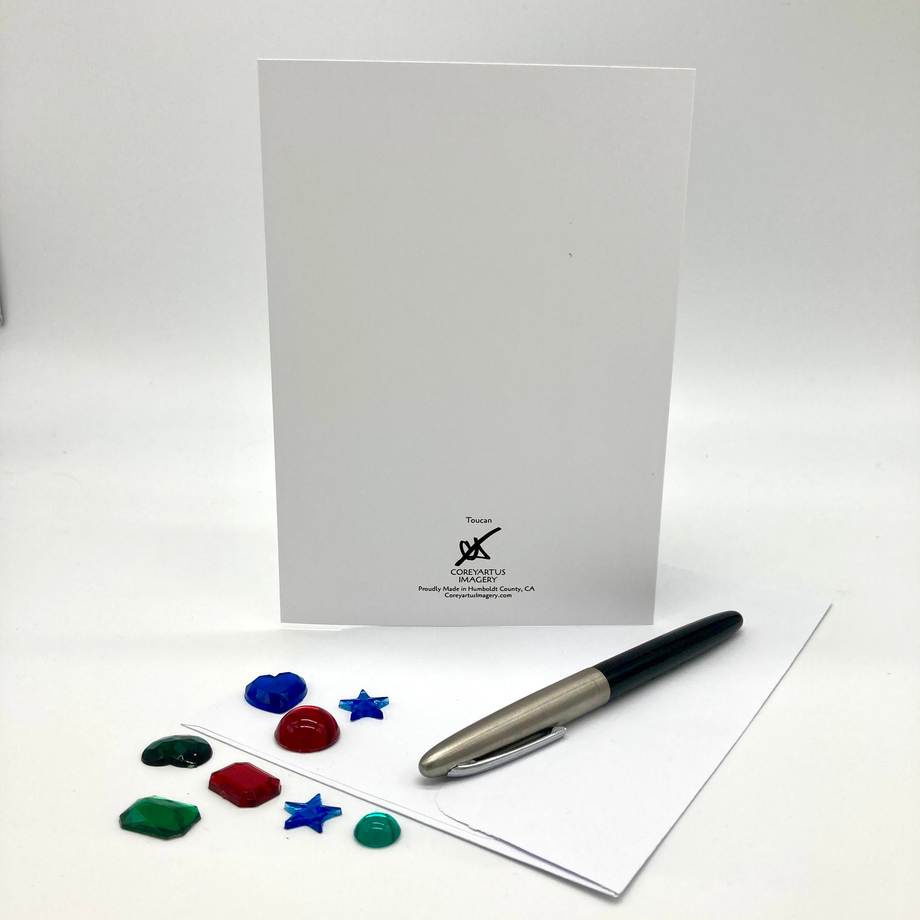 A white stationery set featuring a vertical white card with a small black printed logo at the bottom, a silver and black pen, and several small colorful decorative pieces on a white surface.