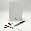 A white stationery set featuring a vertical white card with a small black printed logo at the bottom, a silver and black pen, and several small colorful decorative pieces on a white surface.