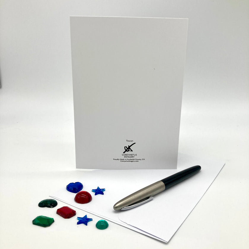 A white stationery set featuring a vertical white card with a small black printed logo at the bottom, a silver and black pen, and several small colorful decorative pieces on a white surface.