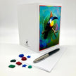 A white greeting card set with a colorful toucan illustration on the front, shown with a pen and small colorful card embellishments on a white background.