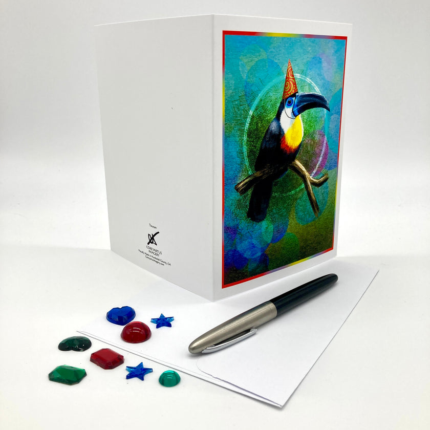 A white greeting card set with a colorful toucan illustration on the front, shown with a pen and small colorful card embellishments on a white background.