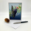 A vertical greeting card featuring a detailed illustration of a woodpecker on a tree trunk against a blue and green bokeh background. The card is displayed standing upright on a white surface next to a pinecone and a black and silver pen.