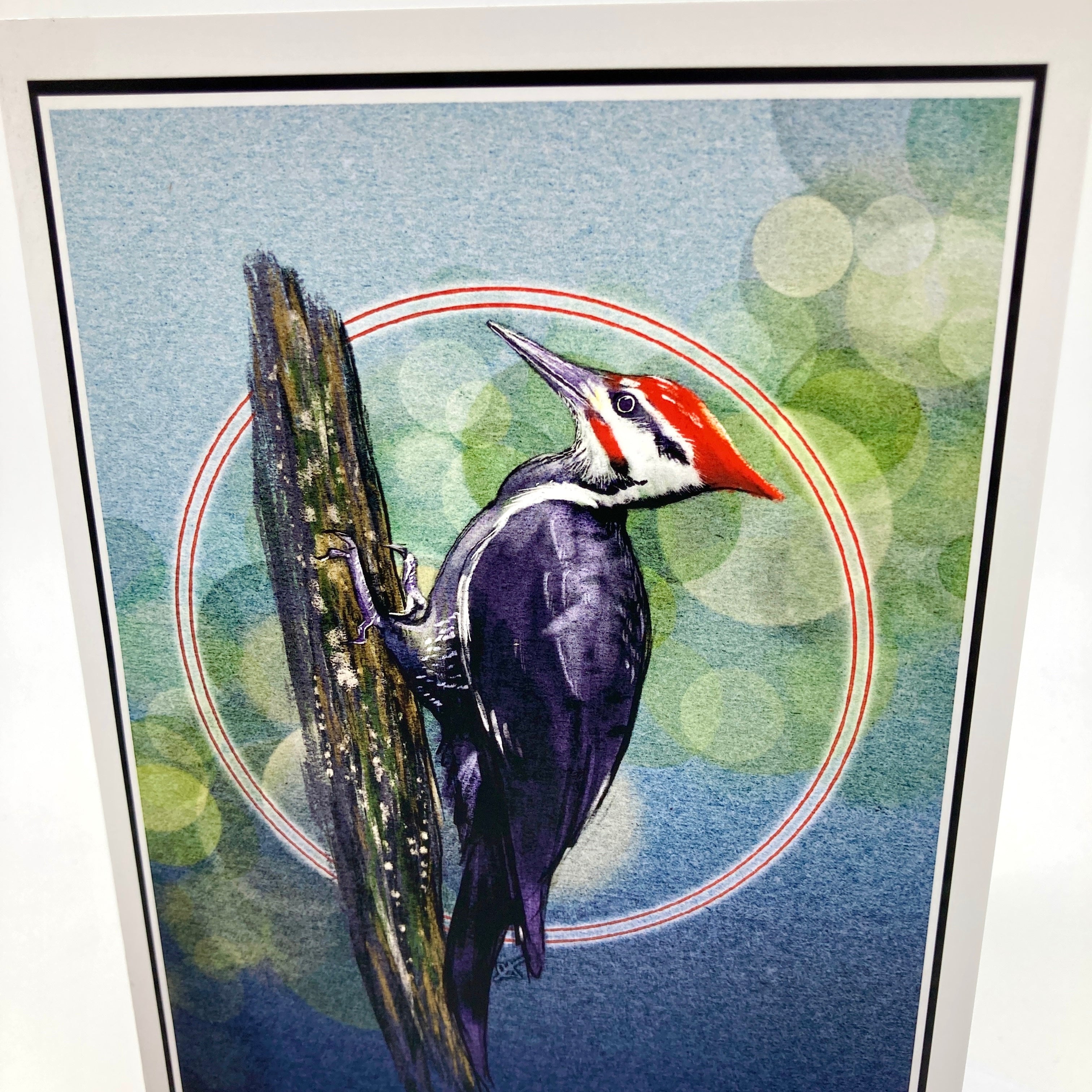 A framed art print featuring a detailed illustration of a woodpecker clinging to a tree trunk, set against a blue and green bokeh background with red circular accents.