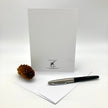A white stationery set featuring a blank white card with black printed branding at the bottom, a white envelope, and a black and silver pen, arranged on a light gray background with a small pinecone prop.