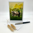 Product photo of an illustration greeting card featuring a mallard duck, posed against a white background and a sprig of grasses and an ink pen in the foreground.