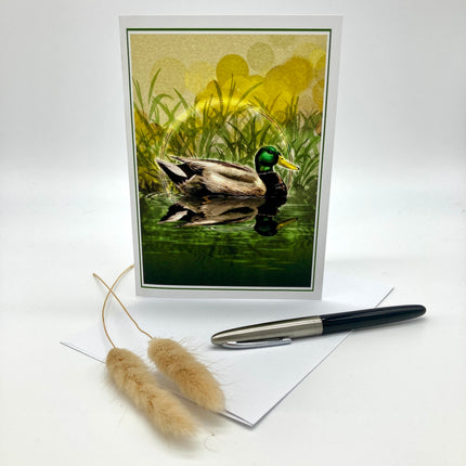 Product photo of an illustration greeting card featuring a mallard duck, posed against a white background and a sprig of grasses and an ink pen in the foreground.