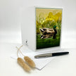 An illustrated greeting card featuring an illustration of a mallard duck, splayed out to show both the front and the back. It is posed against a white background with a grass sprig and an ink pen as accompanying props.