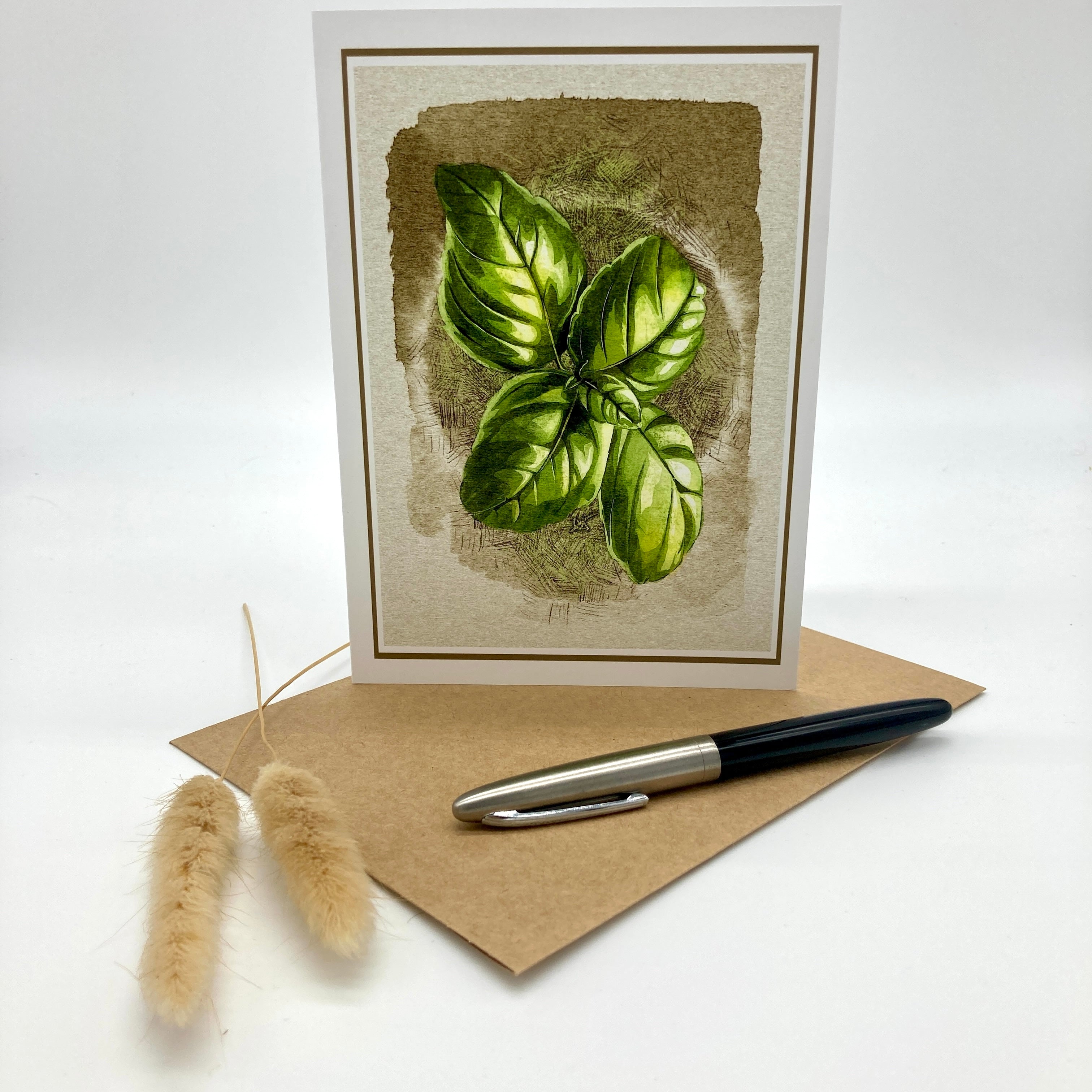 A botanical greeting card featuring a green basil leaf illustration on a textured beige background, displayed upright with a kraft envelope and a black-and-silver pen on a white studio surface. Two small dried bunny tail stems sit beside the envelope.