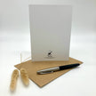 A white greeting card with a small black logo at the bottom stands upright on a brown kraft envelope, with a black and silver pen resting on the envelope and dried grass stems to the left, photographed on a light gray background.