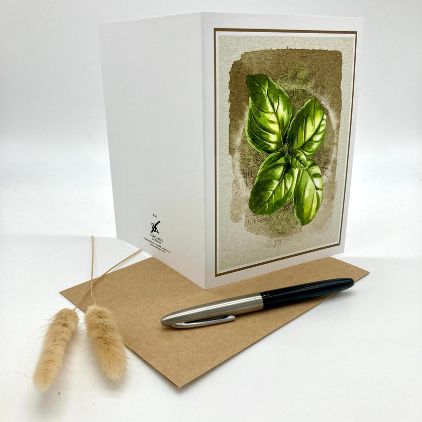 A white greeting card with a botanical illustration of green basil leaves on the front, displayed with a brown kraft envelope and a black-and-silver pen on a white background.