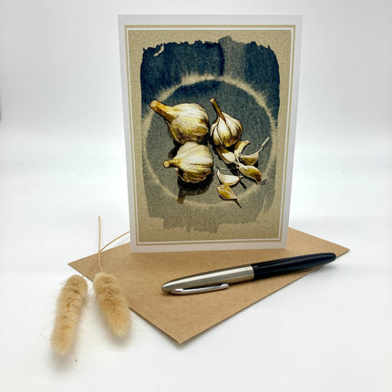 A vertical greeting card featuring a botanical illustration of garlic bulbs, displayed upright on a brown kraft envelope with a black and silver pen and two dried bunny tail stems on a white background.
