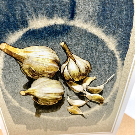 A framed still life art print depicting garlic bulbs and cloves rendered in muted blue-gray, beige, and brown tones.