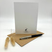A white stationery card with a small black logo at the bottom stands upright on a brown kraft envelope, with a black and silver pen resting on the envelope and two dried bunny tail grass stems to the left, all on a light gray studio background.