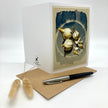 A white greeting card with a botanical illustration of garlic bulbs on the front, displayed with a kraft envelope and a black-and-silver pen on a white background. A small plant sprig decorates the lower left corner.