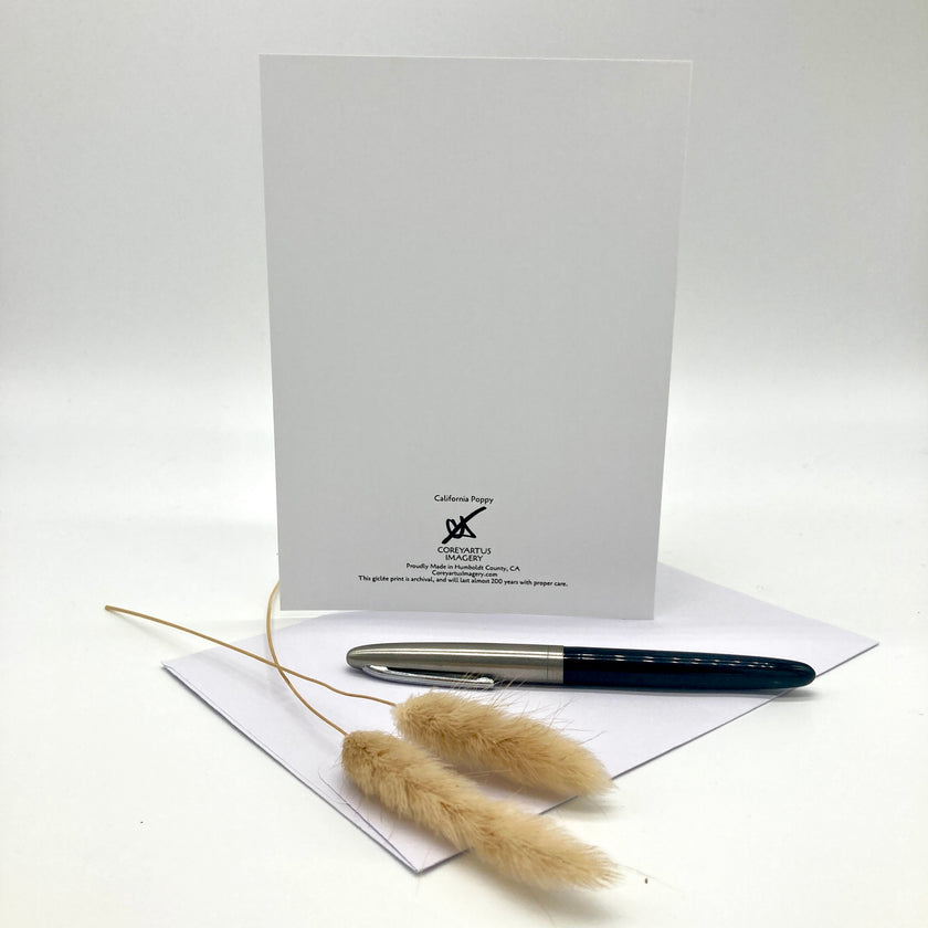 A white greeting card displayed upright with a silver and black pen on white envelopes and dried pampas grass stems in front, against a clean white background.