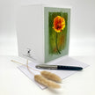 A white greeting card with a printed illustration of an orange-yellow flower on a textured green background, displayed upright with an envelope and a black pen on a white surface. Two dried pampas grass stems rest in front.