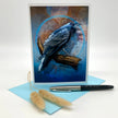 A vertical greeting card featuring a detailed illustration of a black bird perched on a branch against a blue and brown textured background, displayed with a light blue envelope and a black-and-silver pen on a white studio background.