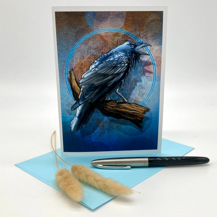 A vertical greeting card featuring a detailed illustration of a black bird perched on a branch against a blue and brown textured background, displayed with a light blue envelope and a black-and-silver pen on a white studio background.
