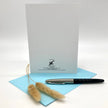 A white card or envelope standing upright on a light blue sheet, with a black and silver pen resting on the blue paper and two dried bunny tail grass stems in front. Small printed branding is visible near the bottom of the white card.