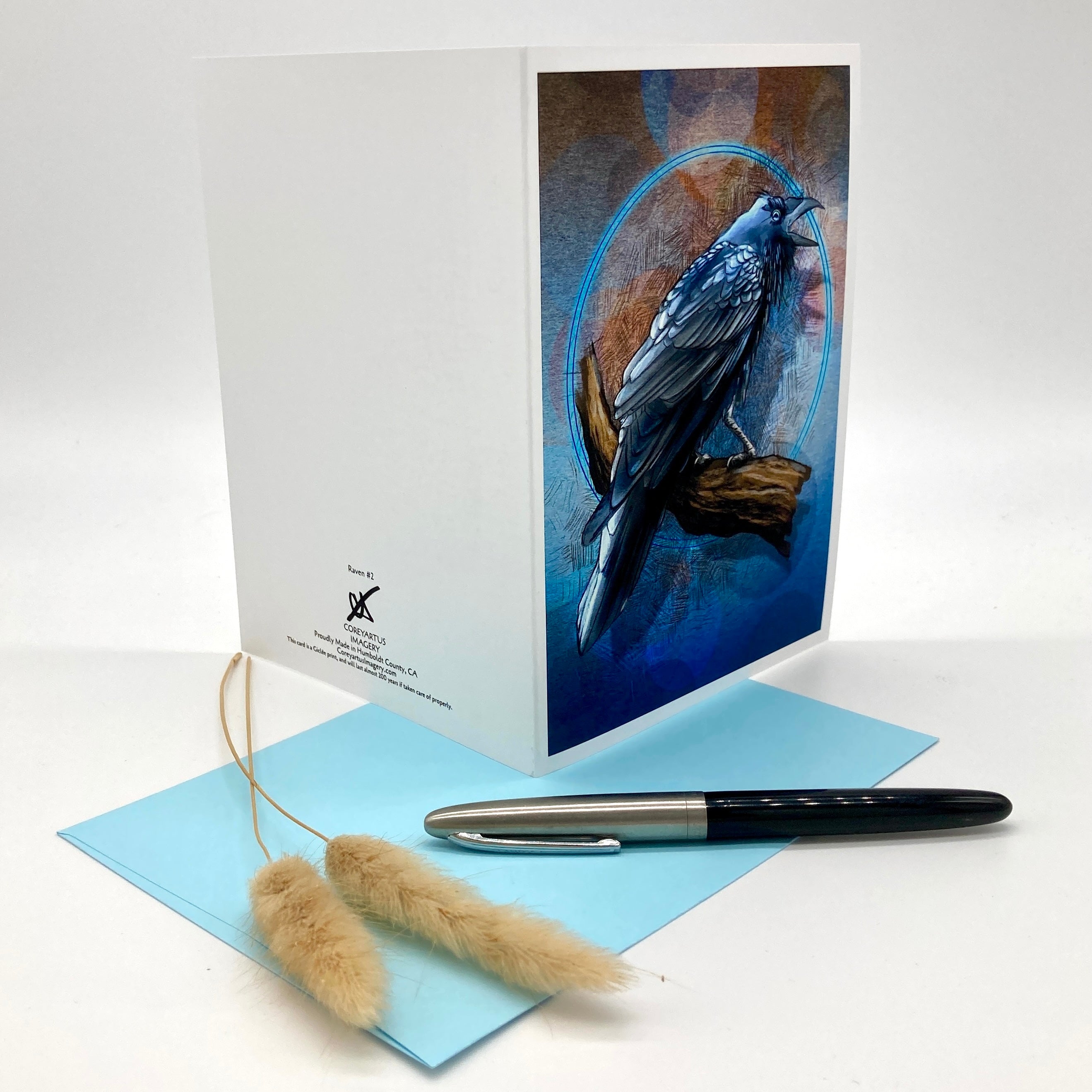 A white greeting card with a detailed illustration of a blue bird on the front, displayed alongside a light blue envelope, a black and silver pen, and two dried bunny tail grass stems on a white background.
