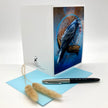 A white greeting card with a detailed illustration of a blue bird on the front, displayed alongside a light blue envelope, a black and silver pen, and two dried bunny tail grass stems on a white background.