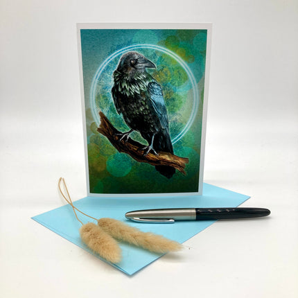 A vertical art print or greeting card featuring a black raven perched on a branch against a teal and green circular background, displayed upright with a light blue envelope, a black and silver pen, and dried pampas grass on a white studio background.