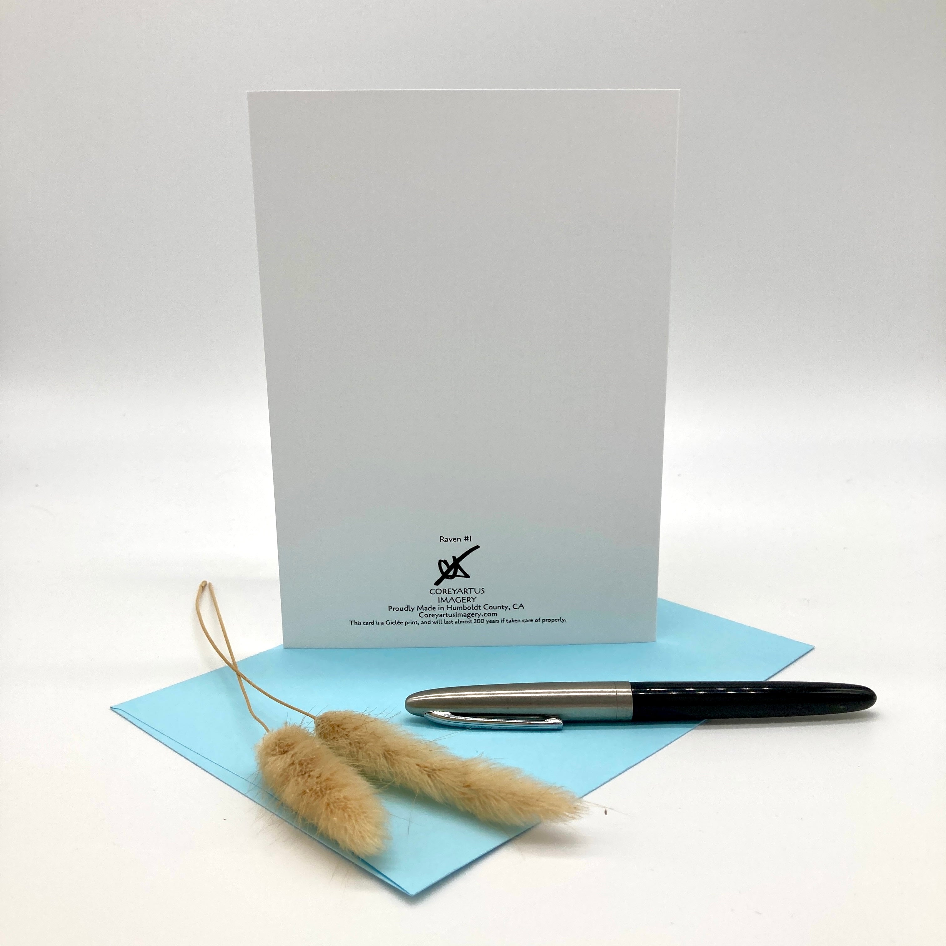 The backside of a greeting card with black printed text and logo, a light blue envelope, and a black and silver pen, styled with dried pampas grass on a white background.