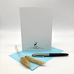 The backside of a greeting card with black printed text and logo, a light blue envelope, and a black and silver pen, styled with dried pampas grass on a white background.