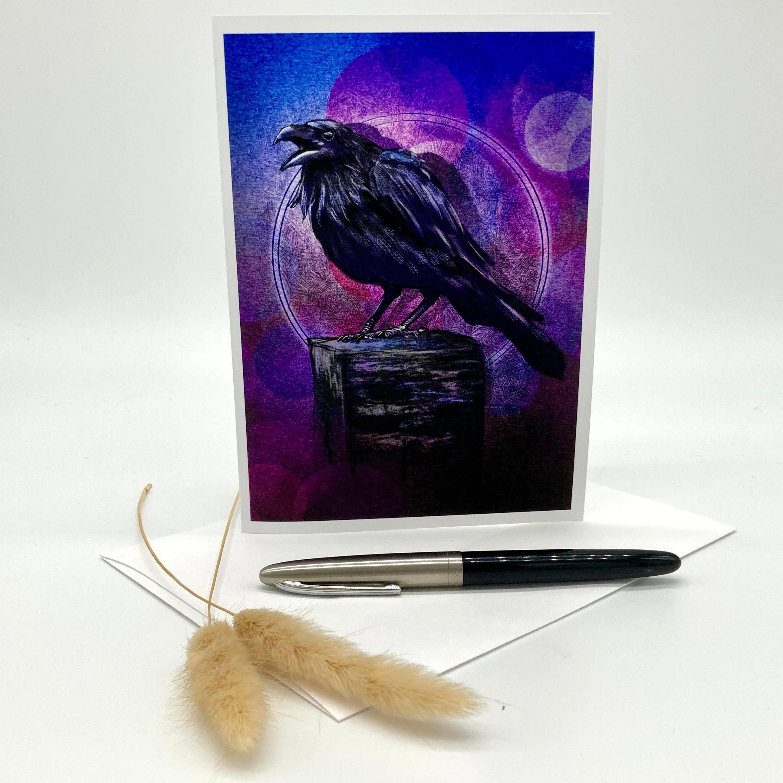 A vertical art print or greeting card featuring a black raven perched on a dark box against a purple and blue abstract background, displayed upright on a white surface with a pen and dried grass stems in front.