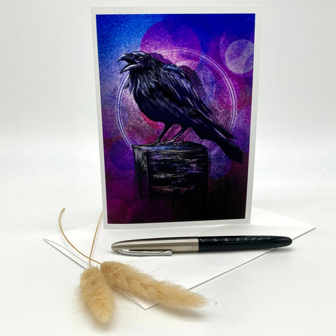 A vertical art print or greeting card featuring a black raven perched on a dark box against a purple and blue abstract background, displayed upright on a white surface with a pen and dried grass stems in front.