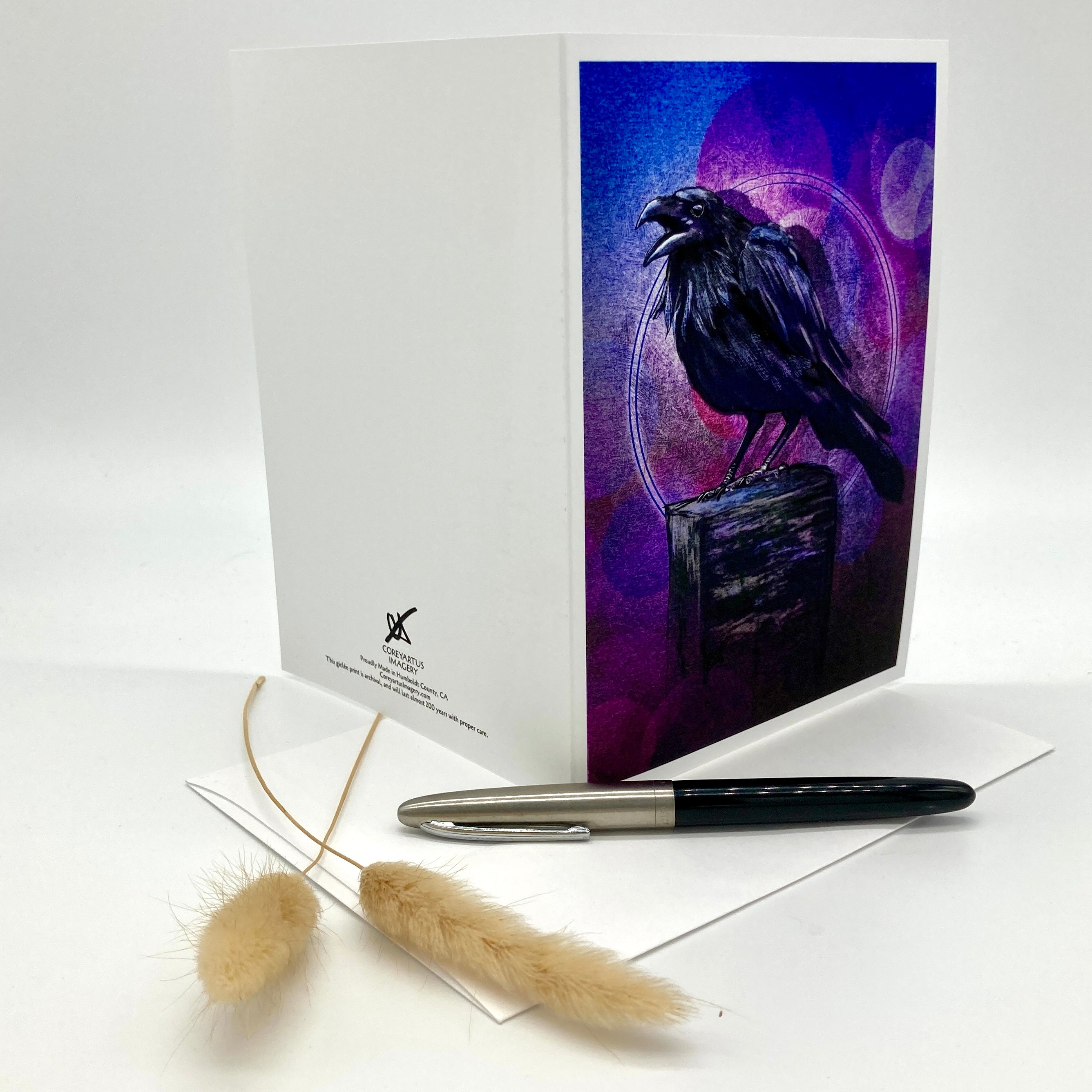 A white greeting card standing upright, showing a printed illustration of a black raven perched on a weathered wooden post against a purple and blue moonlit background. A matching white envelope and a black-and-silver pen lie in front, with dried beige grass stems on a clean white studio background.