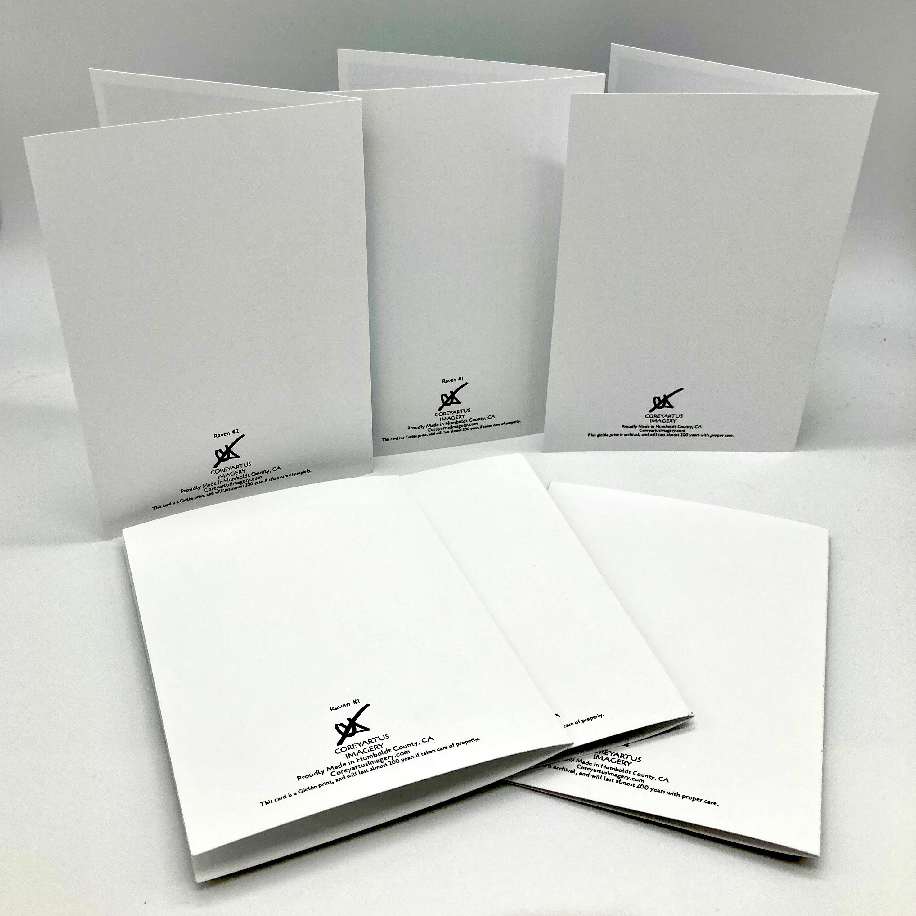 Six white folded paper envelopes or card sleeves arranged on a light gray studio background. Each piece has a small black printed logo and text near the lower edge.