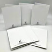 Six white folded paper envelopes or card sleeves arranged on a light gray studio background. Each piece has a small black printed logo and text near the lower edge.