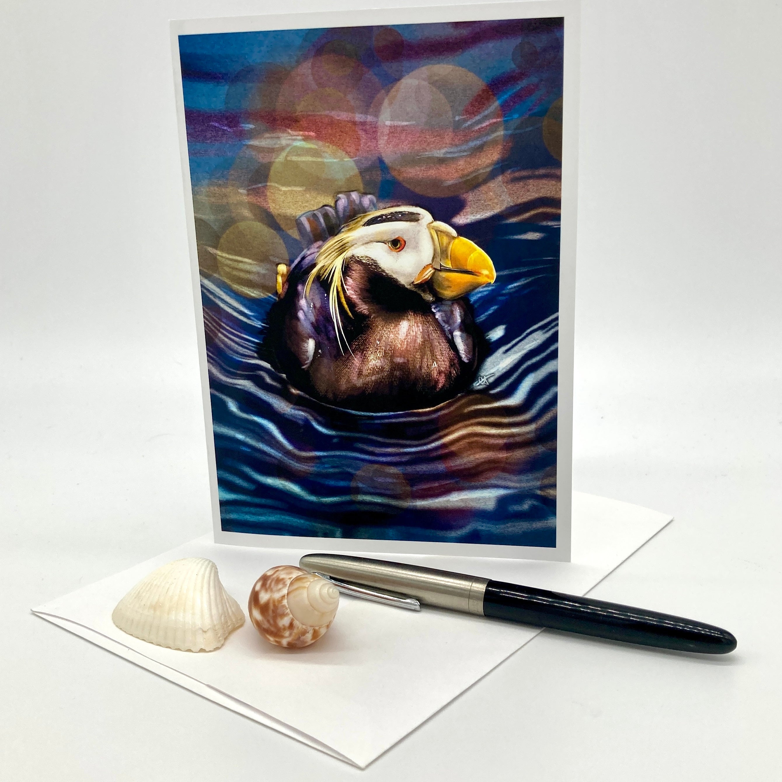 A greeting card featuring a painted puffin in blue water with colorful bokeh light circles, displayed upright on a white surface with a white envelope, a black pen, and two seashells in front.