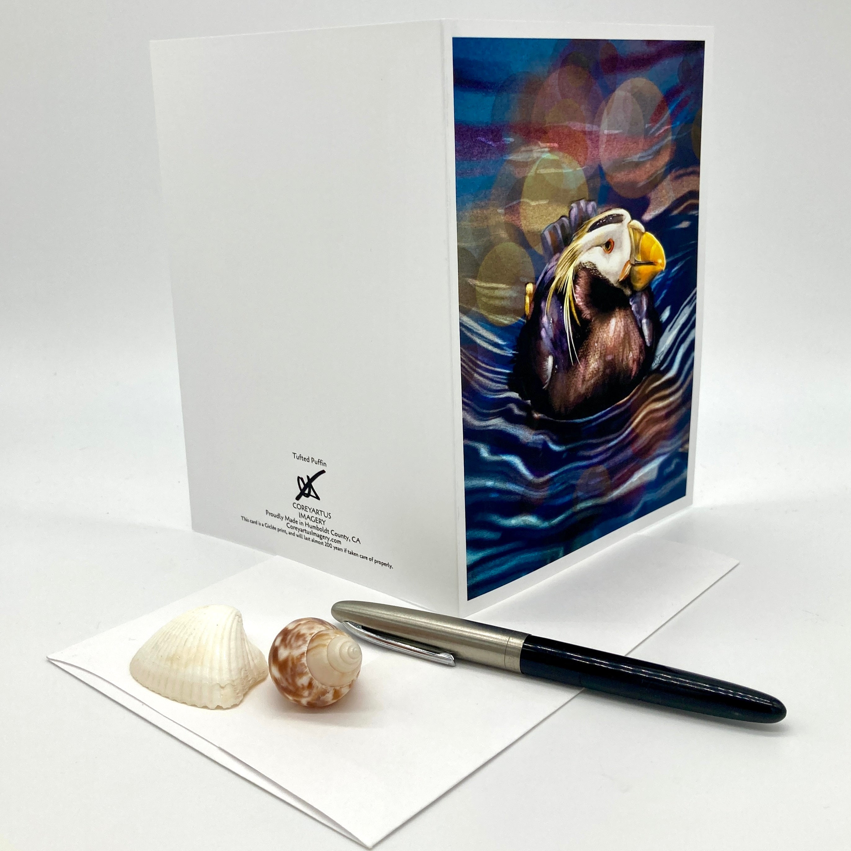 A white greeting card with a colorful puffin illustration on the front, shown standing upright beside an envelope, a black pen, and two seashells on a light gray background.