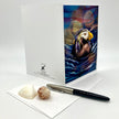A white greeting card with a colorful puffin illustration on the front, shown standing upright beside an envelope, a black pen, and two seashells on a light gray background.