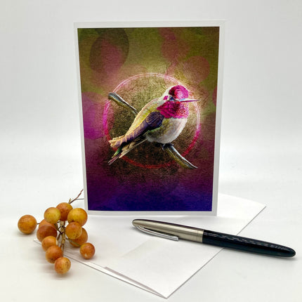 A art print greeting card featuring a hummingbird perched on branches with a glowing circular motif and pink-toned background, styled on a white paper envelope and surface with a white envelope and a black pen nearby.